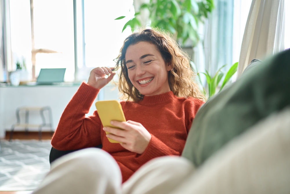 A young woman with curly hair wearing a red sweater, smiling and holding a smartphone while sitting on a couch