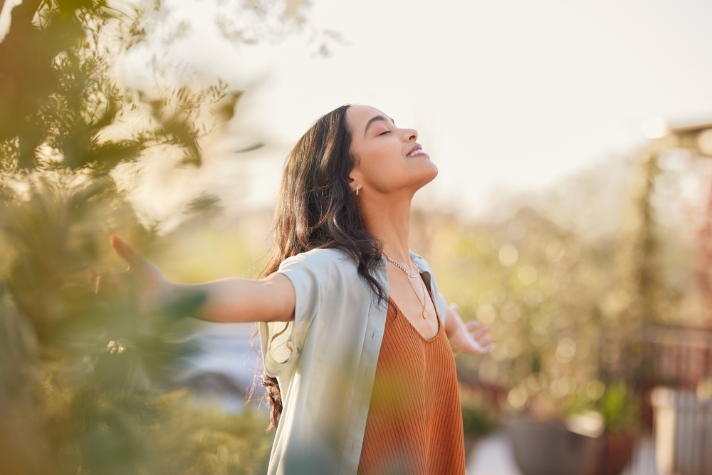 A woman with long brown hair enjoying the outdoors, with her arms outstretched and her eyes closed, surrounded by greenery