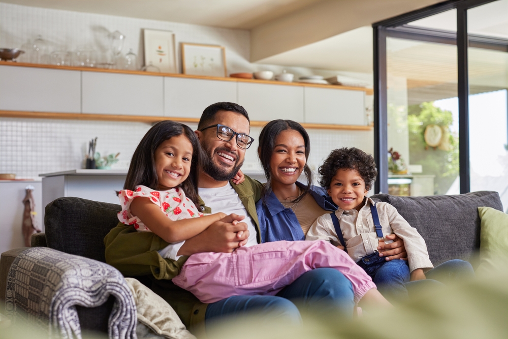 A happy family of four - a mother, father, and two young children - sitting together on a couch in a cozy living room. They are all smiling and appear to be enjoying each other's company.